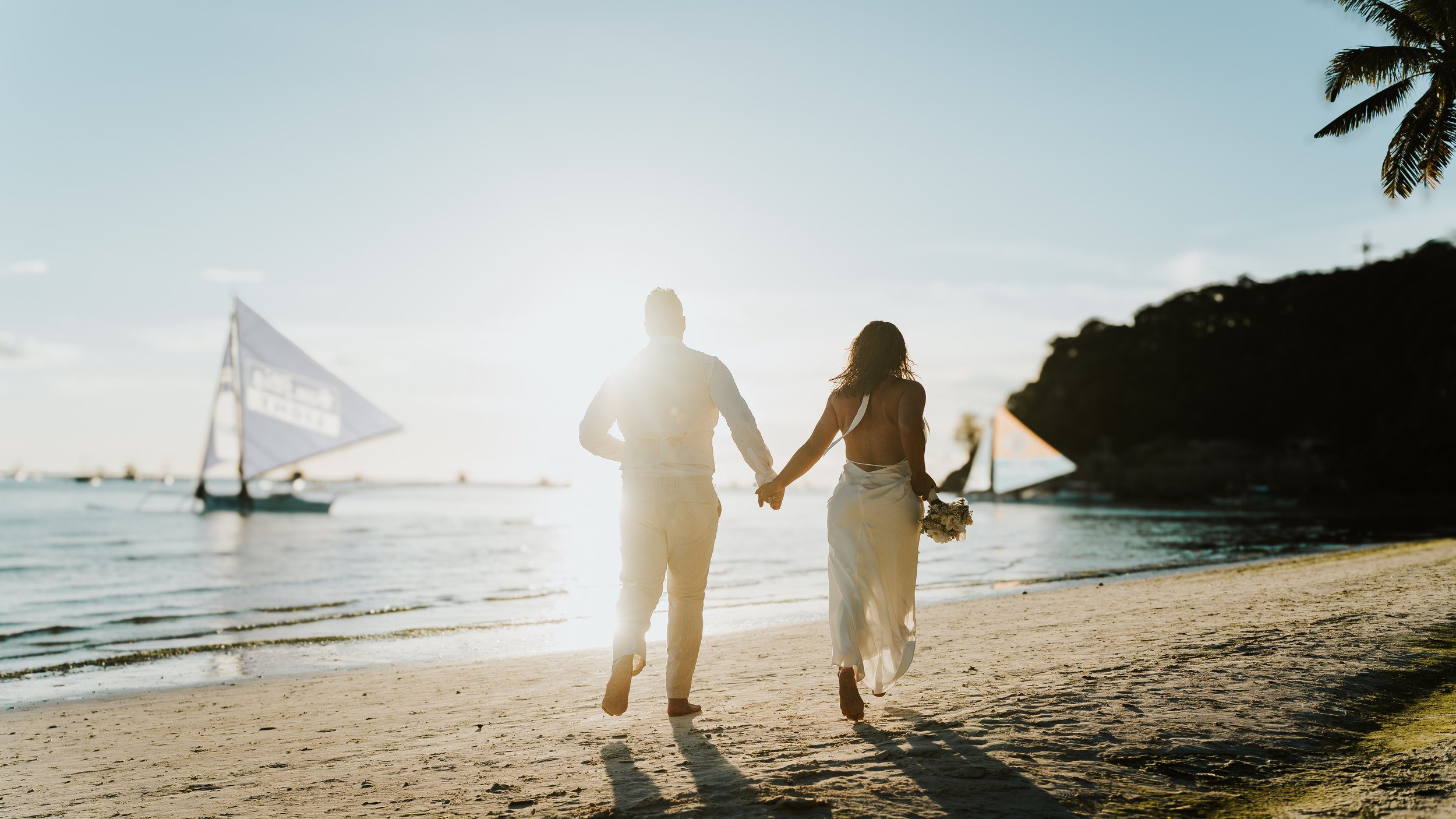 Silhouetted bride and groom holding hands walking on beach at sunset, with sailboats in the water.
