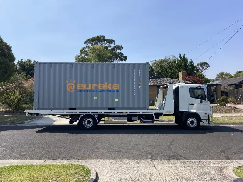 A white flatbed truck carrying a gray shipping container with "Eureka Logistics & Containers" branding on a suburban street.