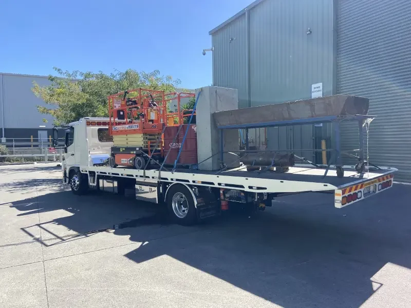 Flatbed truck carrying construction equipment, including scissor lifts, parked outside an industrial building.