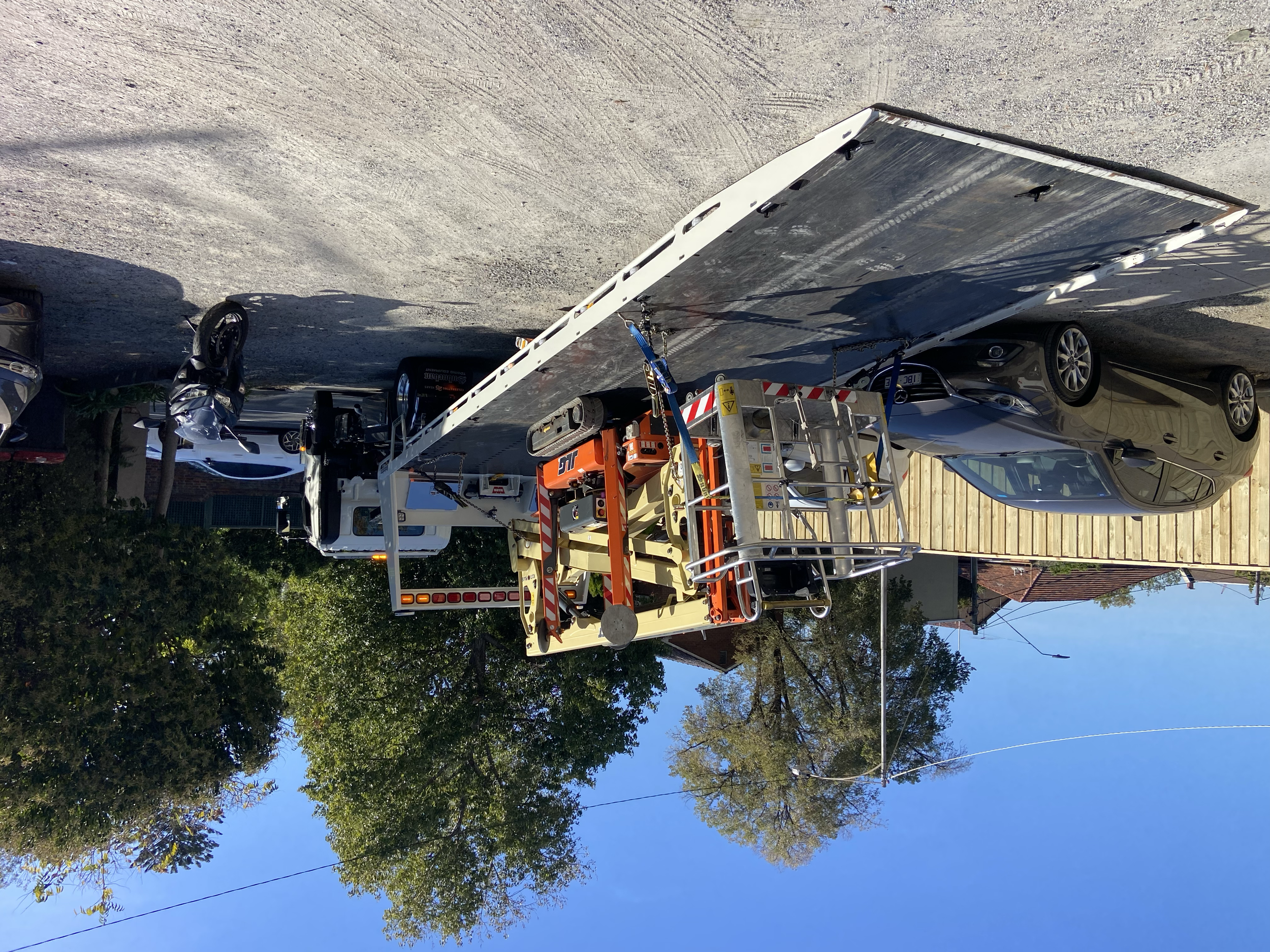 Flatbed truck carrying construction equipment and a motorcycle parked on a street with cars around, trees, and a clear blue sky.