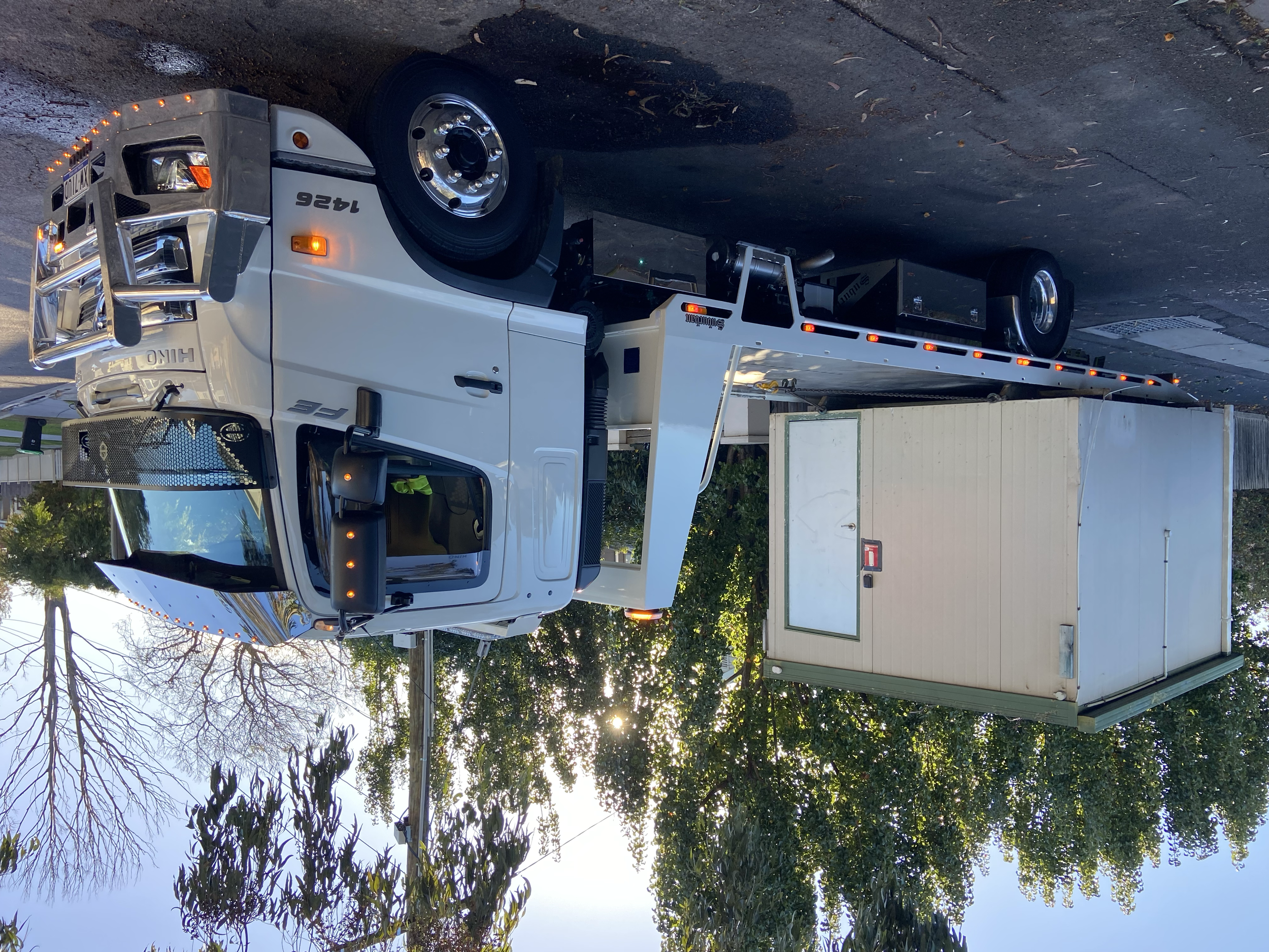 Tow truck idling on street with portable building on back, surrounded by trees.