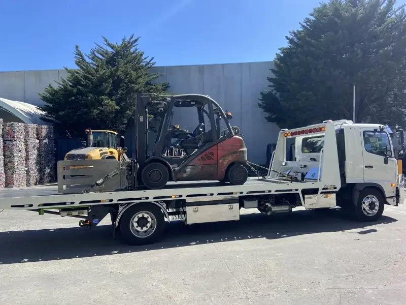 A white tow truck transporting a forklift on its flatbed in an industrial area, with bales of crushed cans and trees in the background.