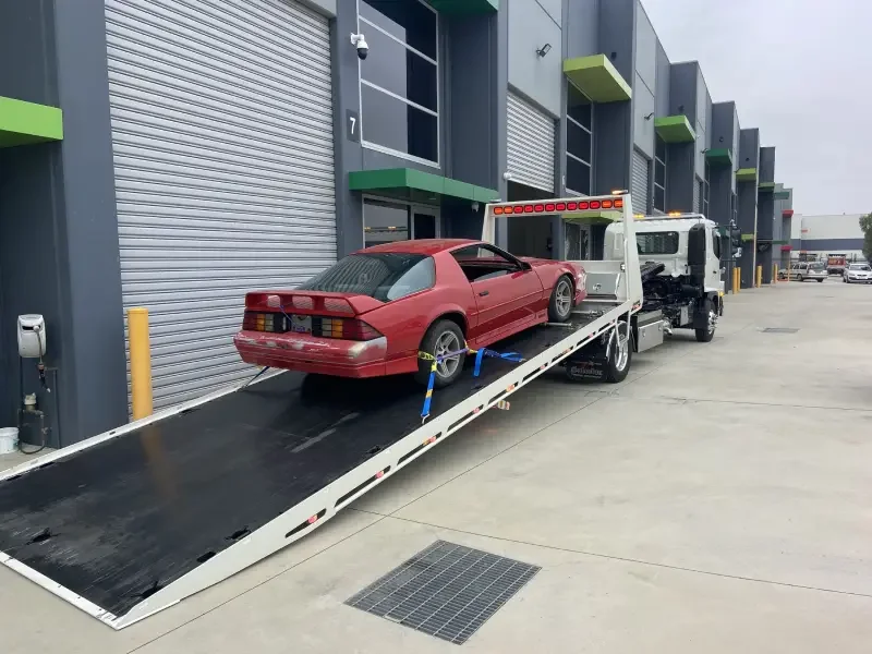 Red sports car on a flatbed tow truck near industrial buildings.