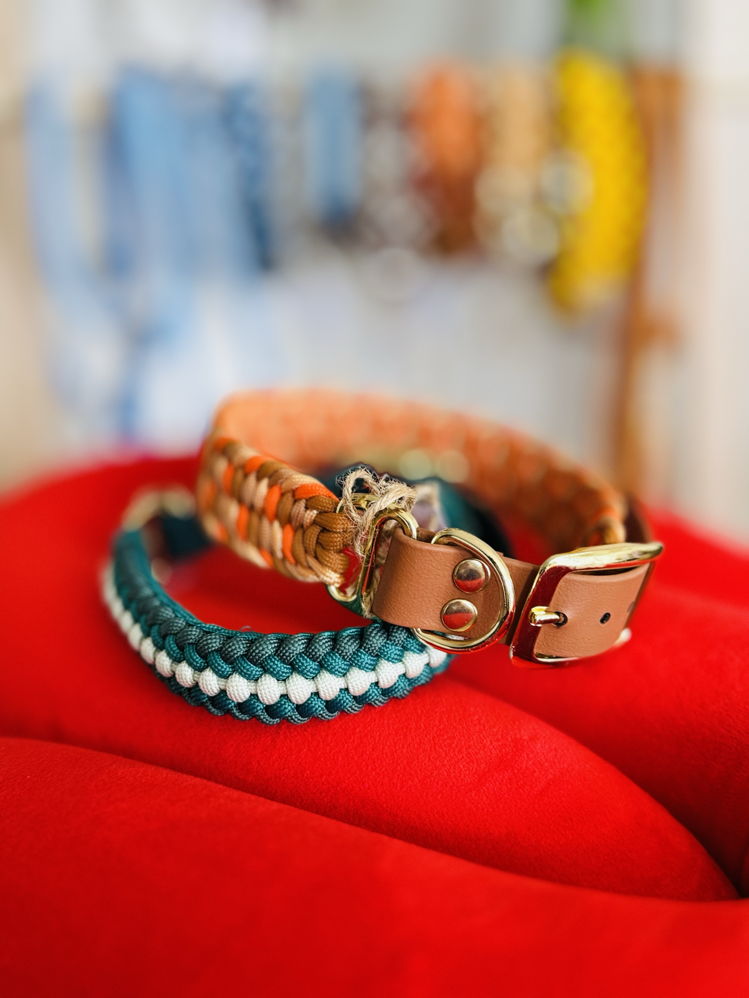 Deux colliers pour chien, un tressé en bleu et blanc, l'autre en orange et beige, posés sur un tabouret rouge. En arrière-plan, des accessoires de chien suspendus à un présentoir en forme d'échelle. Le tout, de la marque belge "Comme Cha et chiens".