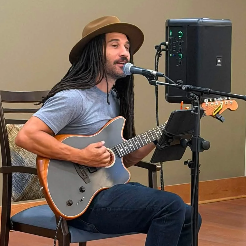 Man with long dreadlocks, wearing a tan hat and gray t-shirt, playing an acoustic guitar and singing into a microphone in a small indoor space.