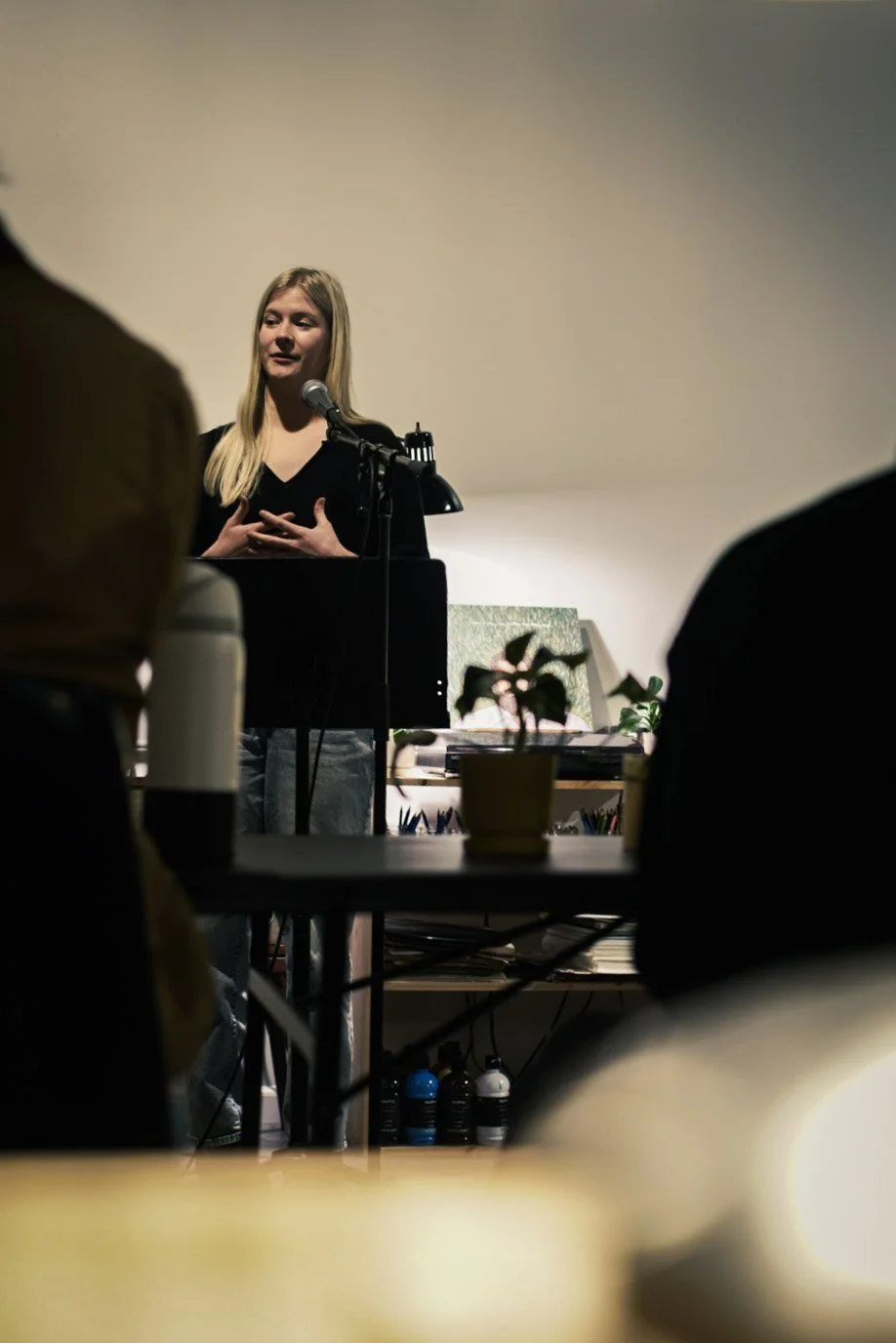 A woman with long blonde hair standing at a podium speaking into a microphone about her poetry, with several audience members in the foreground, in an indoor setting with potted plants and various items on a table behind her.