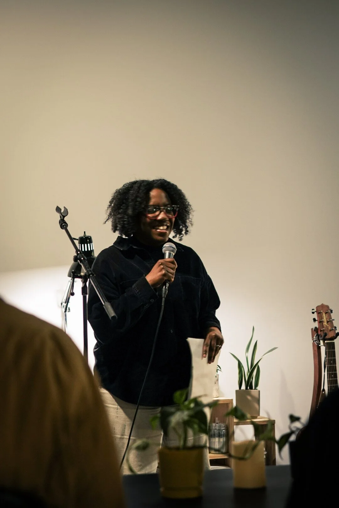 A woman with glasses and curly hair speaking into a microphone at an indoor event called Field Co Nite about her journey as an artist. There is a guitar, potted plants, and audience members visible in the foreground.