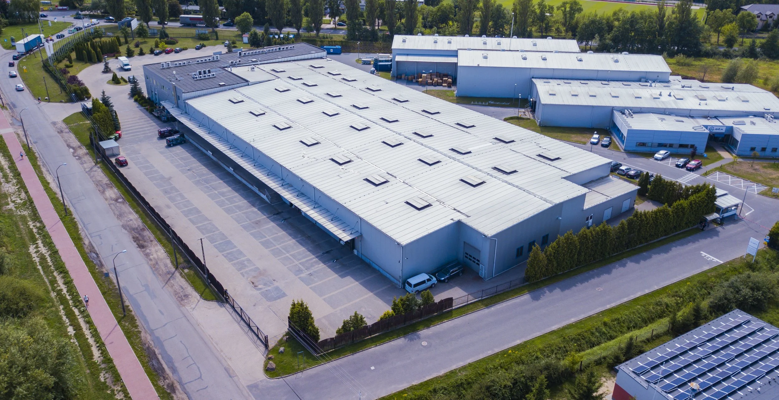 Aerial view of an industrial warehouse complex with parking lot, surrounded by trees and greenery.