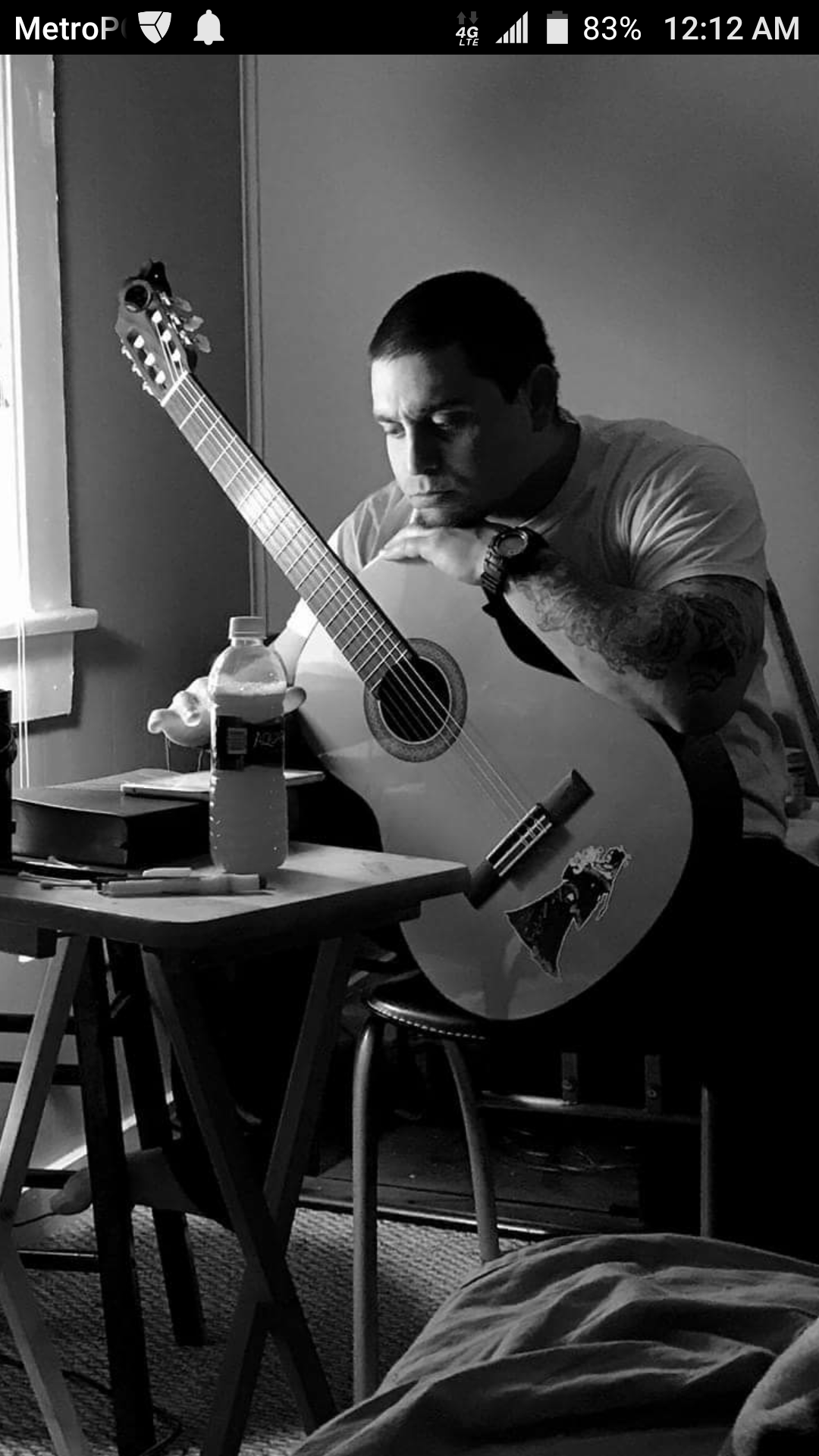 A black and white photograph of a man sitting at a small table with a guitar, resting his chin on his folded arms, and looking thoughtful. The table has a water bottle, some papers, and a few pens. The room has a window with blinds, a wall, and a chair. The man is wearing a wristwatch and has tattoos on his arm.