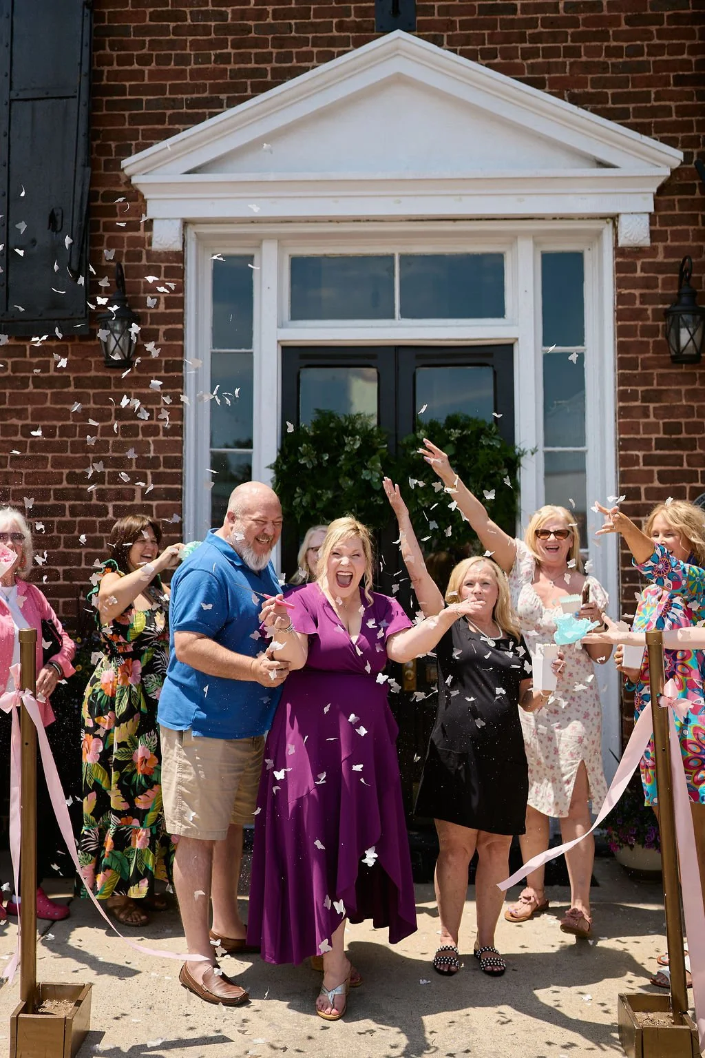 People celebrating outside a brick building with paper confetti falling around them. They are smiling, cheering, and throwing confetti during a festive gathering.