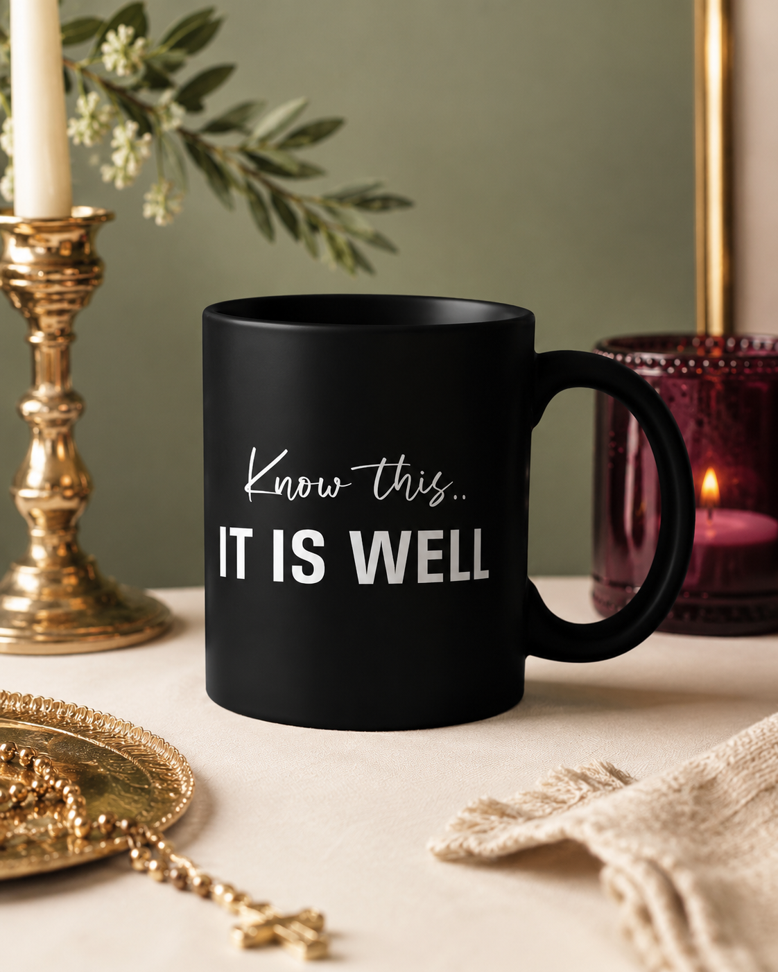 A black mug with the words "Know this... It is well" written on it, placed on a white surface alongside gold and purple decorative items and green foliage.