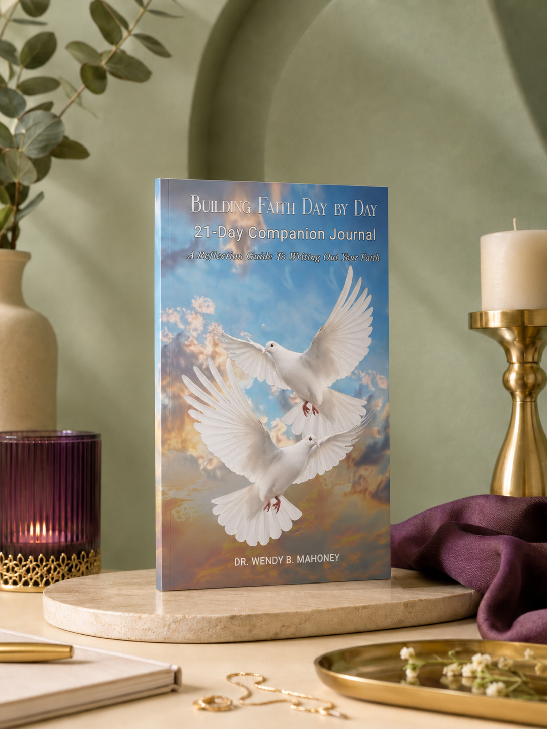 Book titled "Building Faith Day by Day: 21-Day Companion Journal" with two white doves flying against a sky background. The scene includes a purple candle holder, a gold candle holder with a white candle, a purple cloth, a closed notebook, gold jewelry, a gold tray, and small white flowers on a beige surface with a green wall in the background.