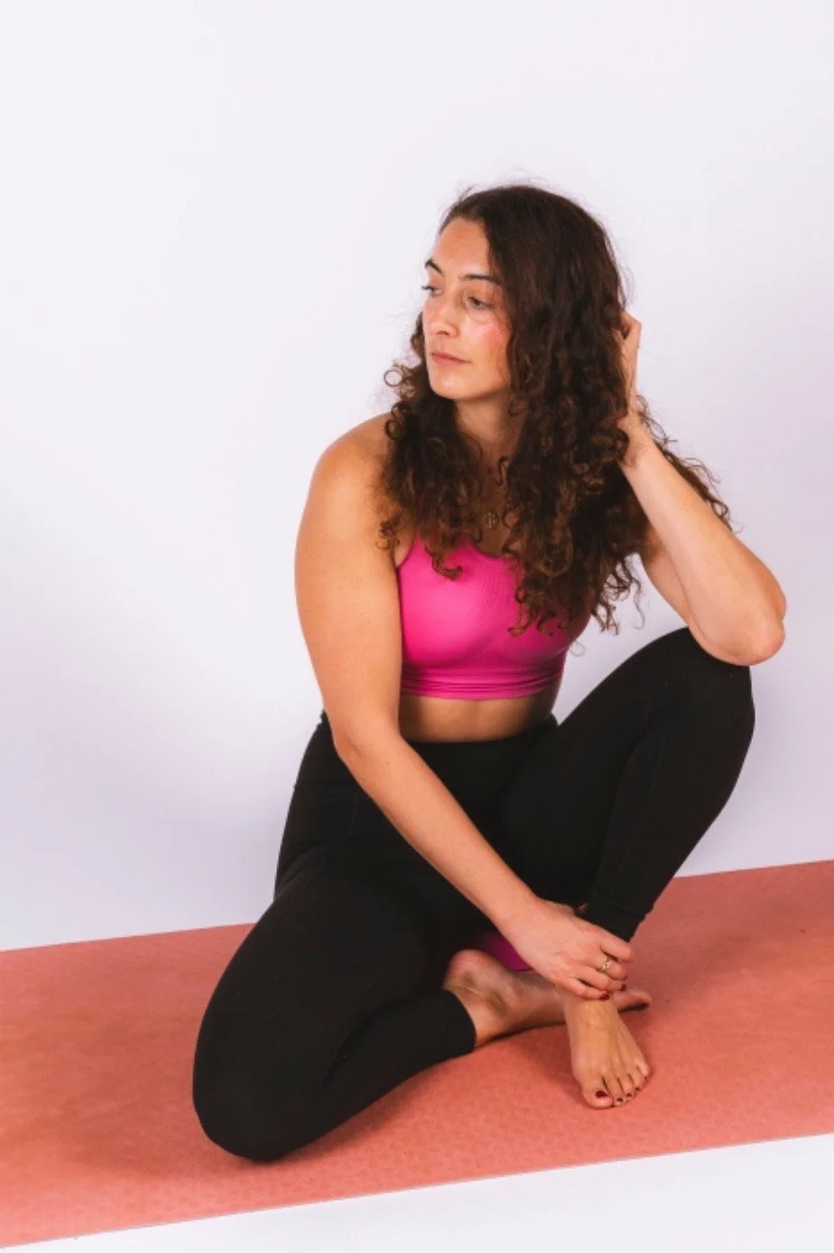 A woman with curly brown hair practicing yoga, wearing a pink sports bra and black leggings on a pink yoga mat against a white background.