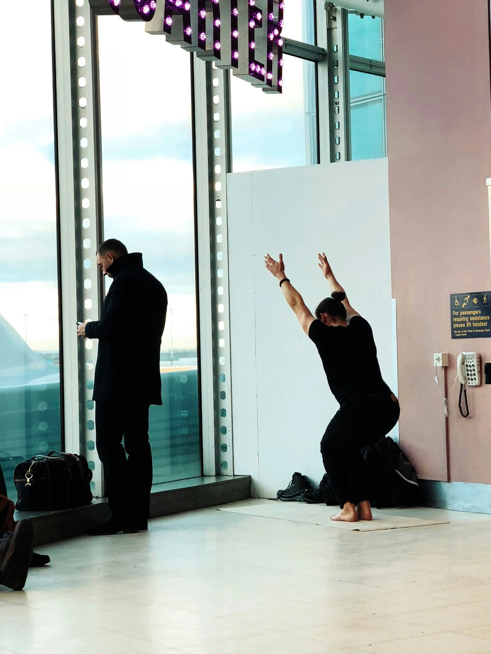 A woman with her hair tied back is kneeling on a prayer mat, raising her arms in prayer at an airport terminal near large windows. A man is standing nearby, looking at his phone, with bags placed on the floor.
