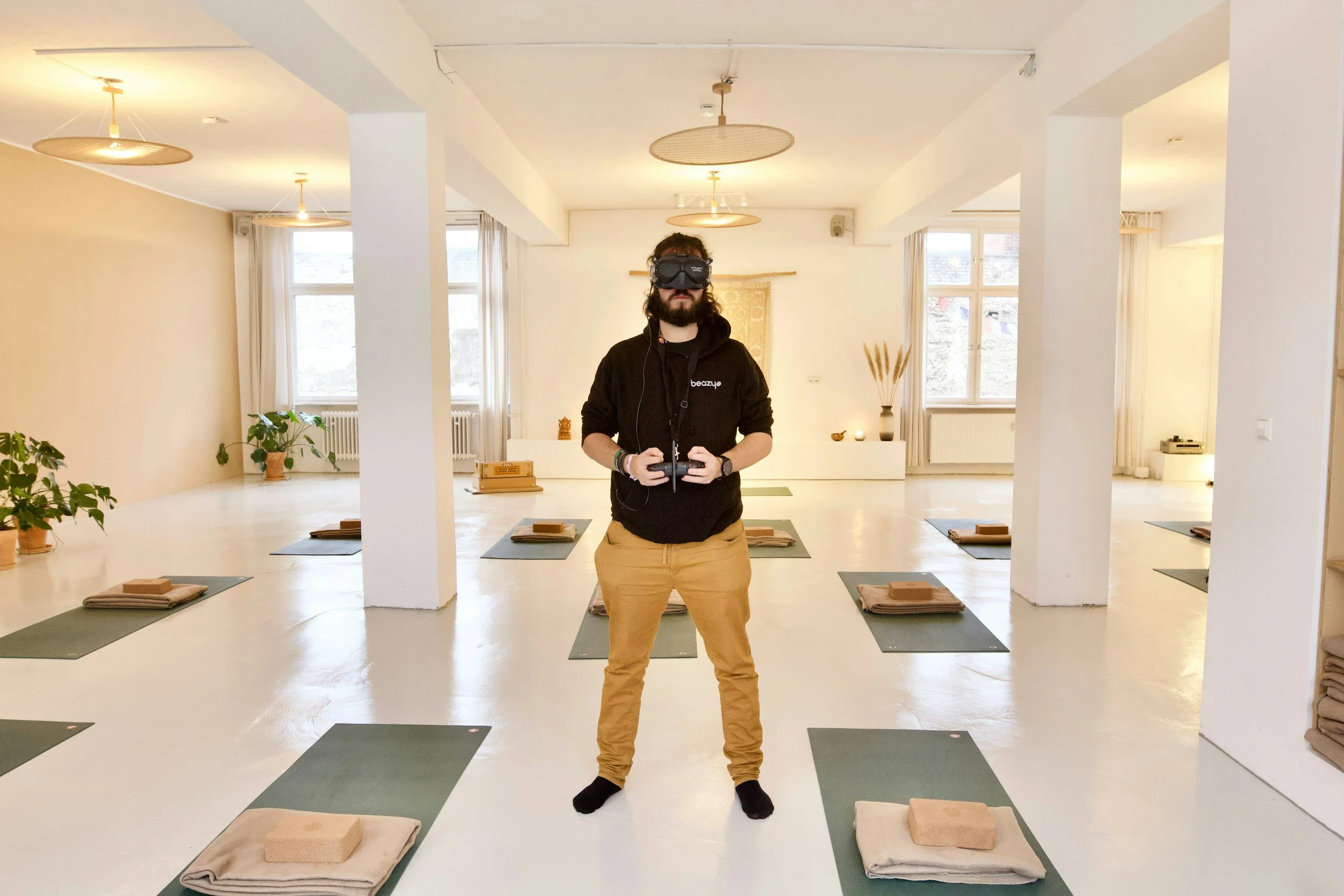 Man wearing virtual reality goggles holding a controller in a minimalist yoga studio with mats, towels, and bricks on the floor