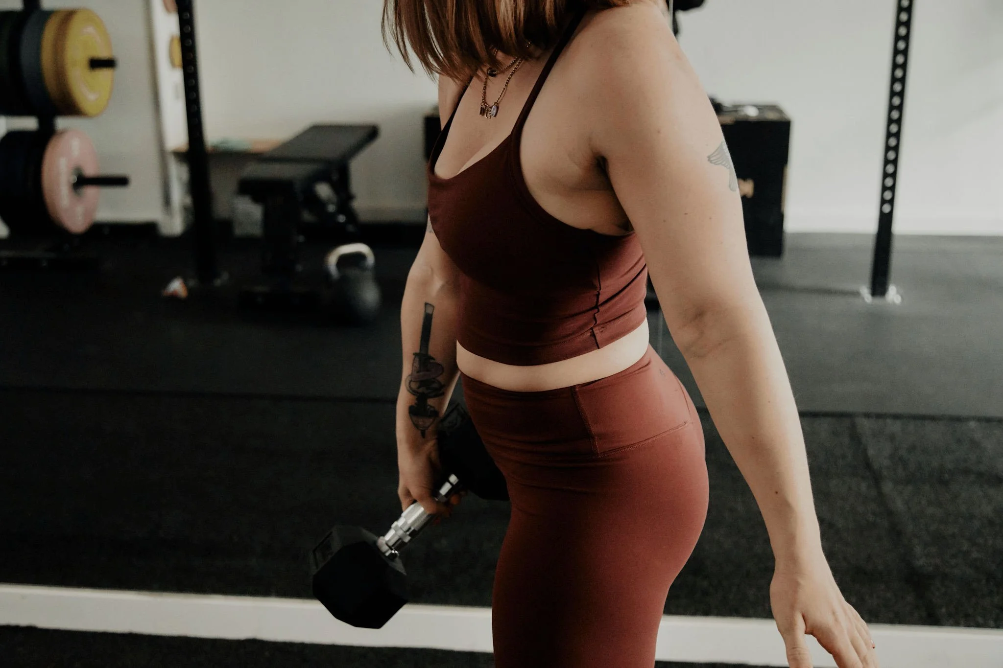Eine Frau hebt in einem Fitnessstudio eine Hantel. Sie trägt braune Sportkleidung und hat Tattoos auf ihrem Arm.