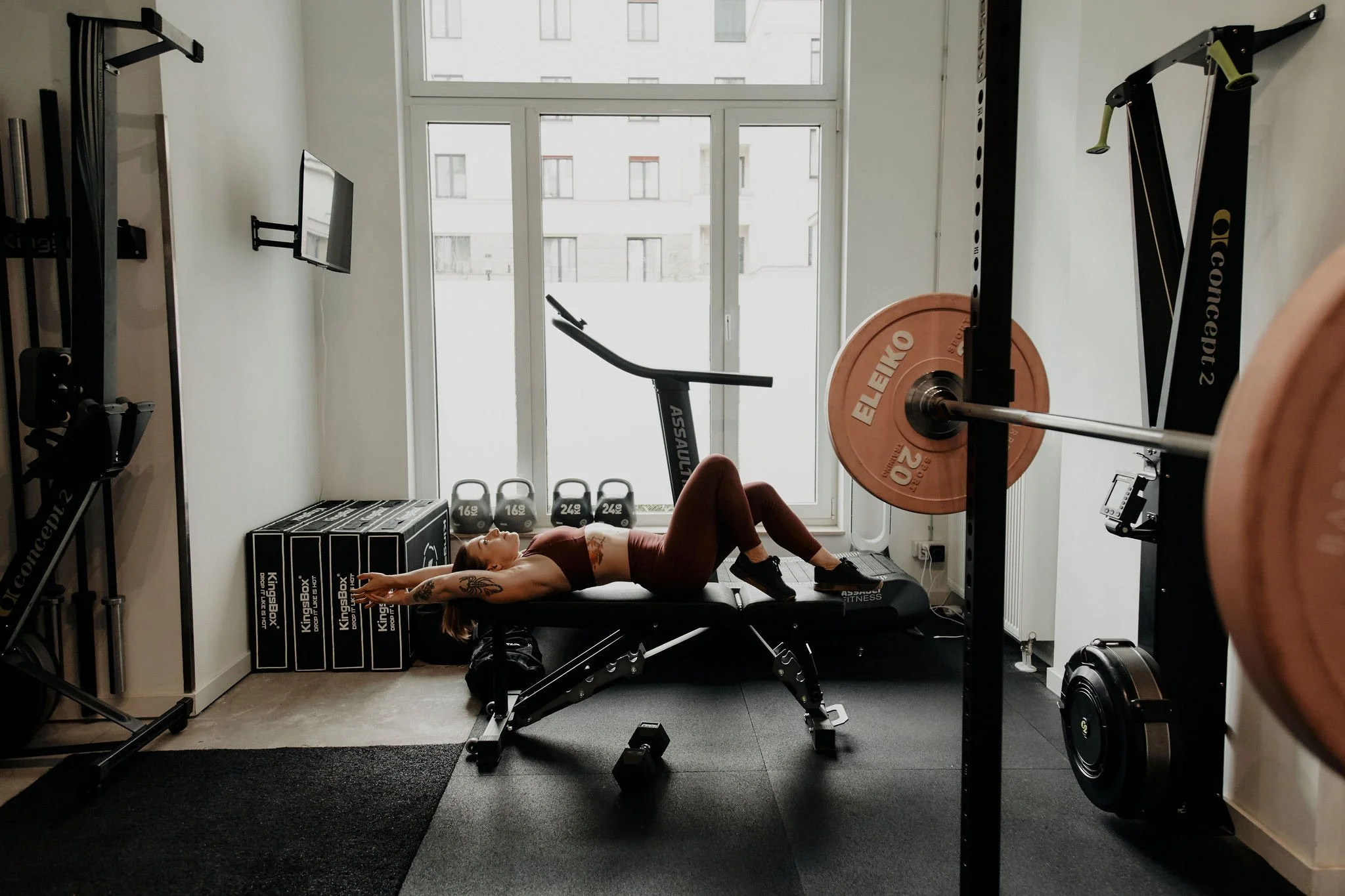 Frau liegt auf einer Trainingsbank im Fitnessstudio, im Hintergrund eine große Fensterfront, in der Gym-Ausrüstung wie Hanteln, Ganzkörpertrainingsgerät und ein Fernseher zu sehen sind.