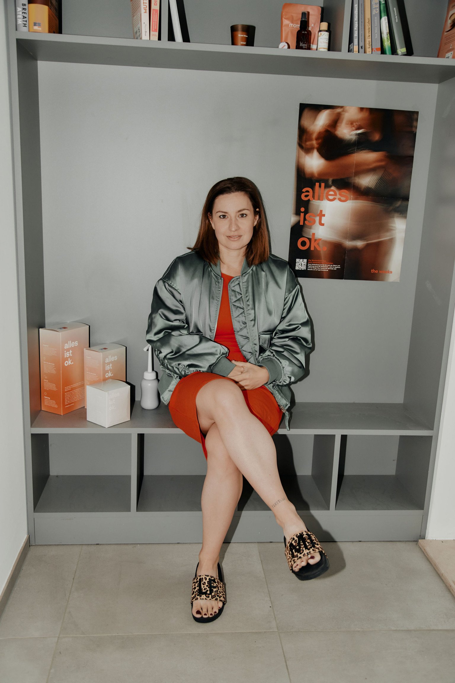 Eine Frau mit braunen Haaren sitzt auf einer grauen Sitzecke, trägt ein rotes Kleid, eine graue Jacke und leopardmusterte Hausschuhe, vor einem grauen Regal mit Büchern und Produkten, neben einem Poster mit dem Text "alles ist ok."