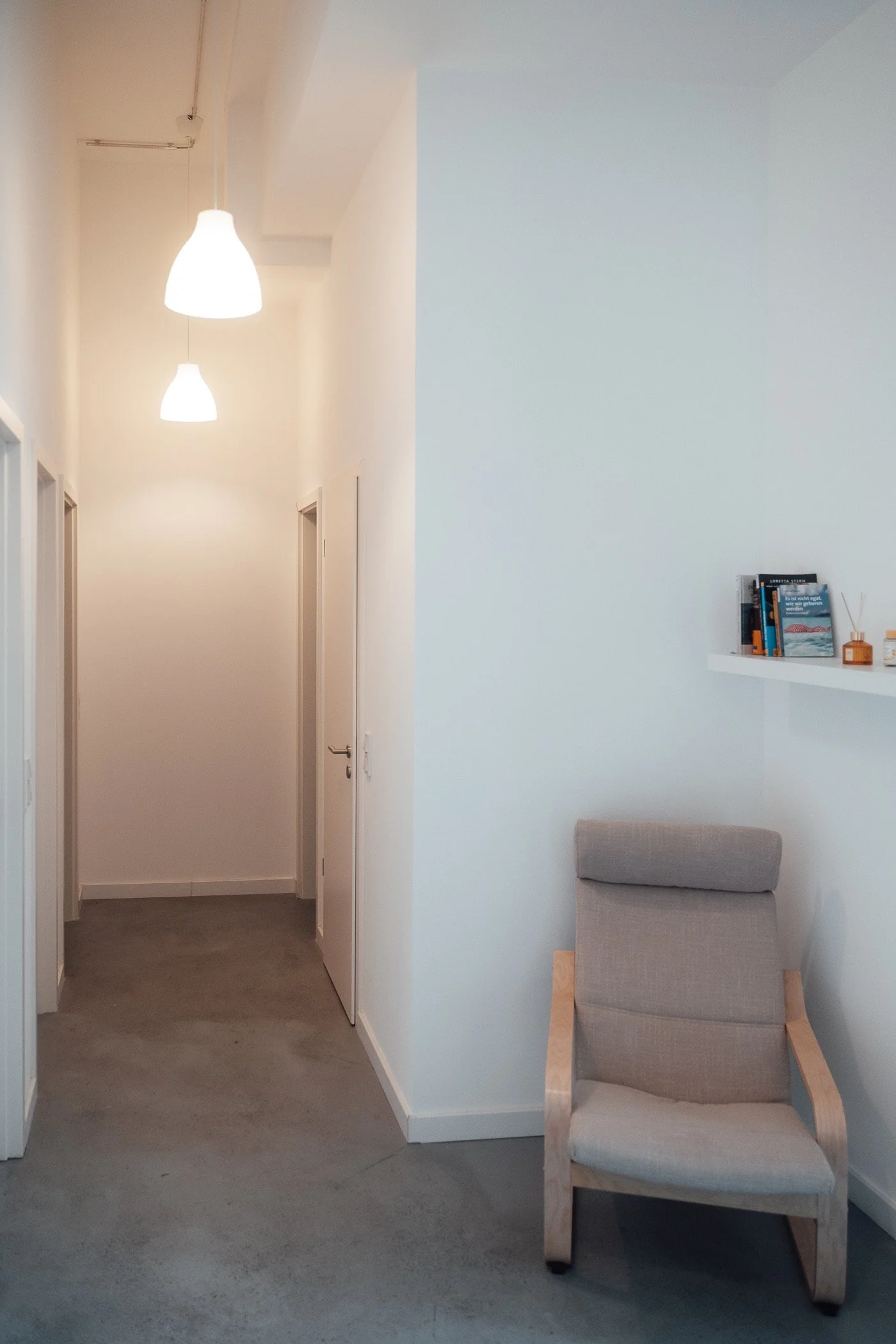 Ein Flur mit weißen Wänden, Deckenleuchten und einem beigen Sessel am rechten Rand, obere Regale mit Büchern und Dekorationsgegenständen.