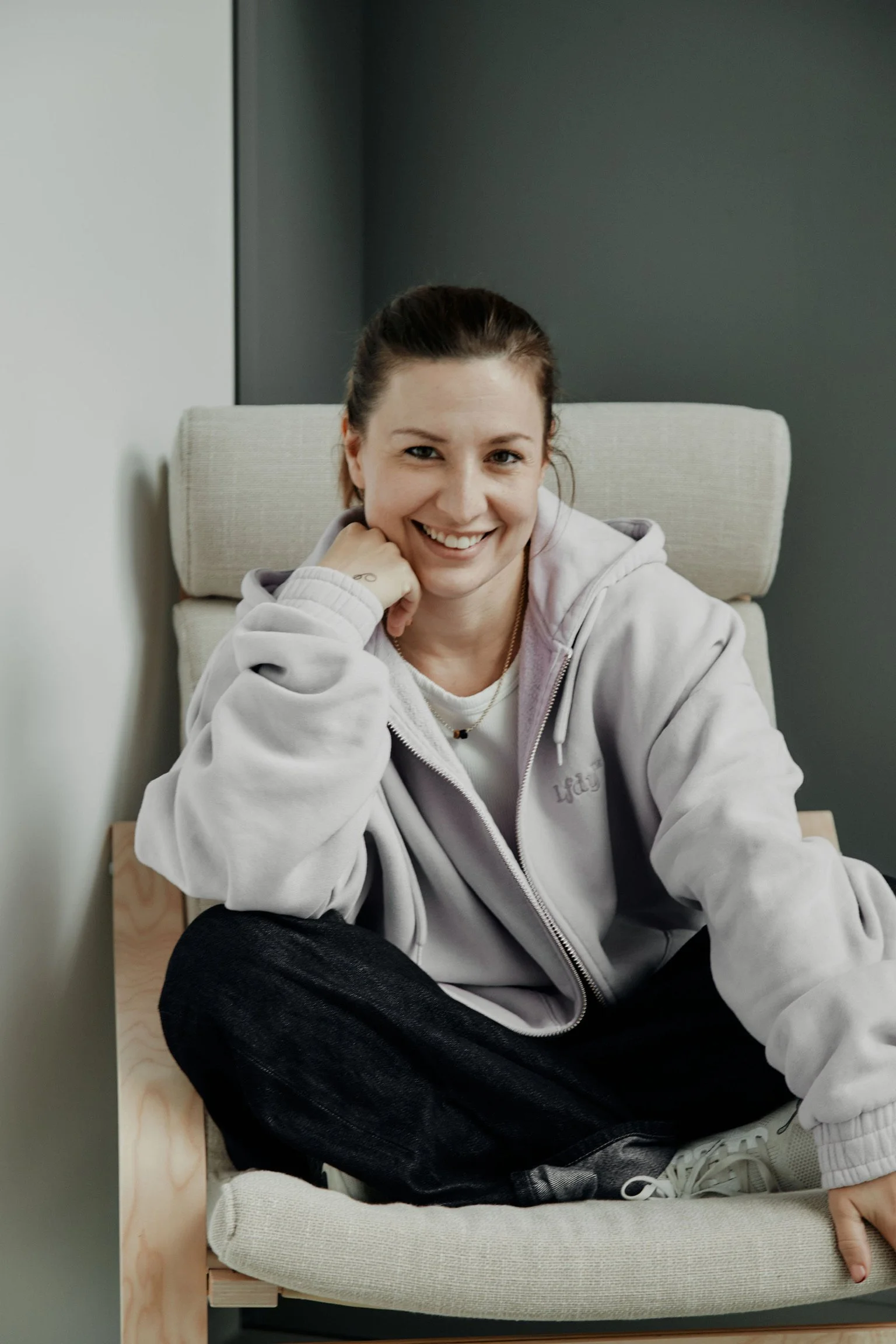 Junge Frau mit braunen Haaren, smile, schwarze Haare, trägt graue Kapuzenjacke, sitzt auf einem Stuhl, Sofa, lächelnd, casual Kleidung, Indoor