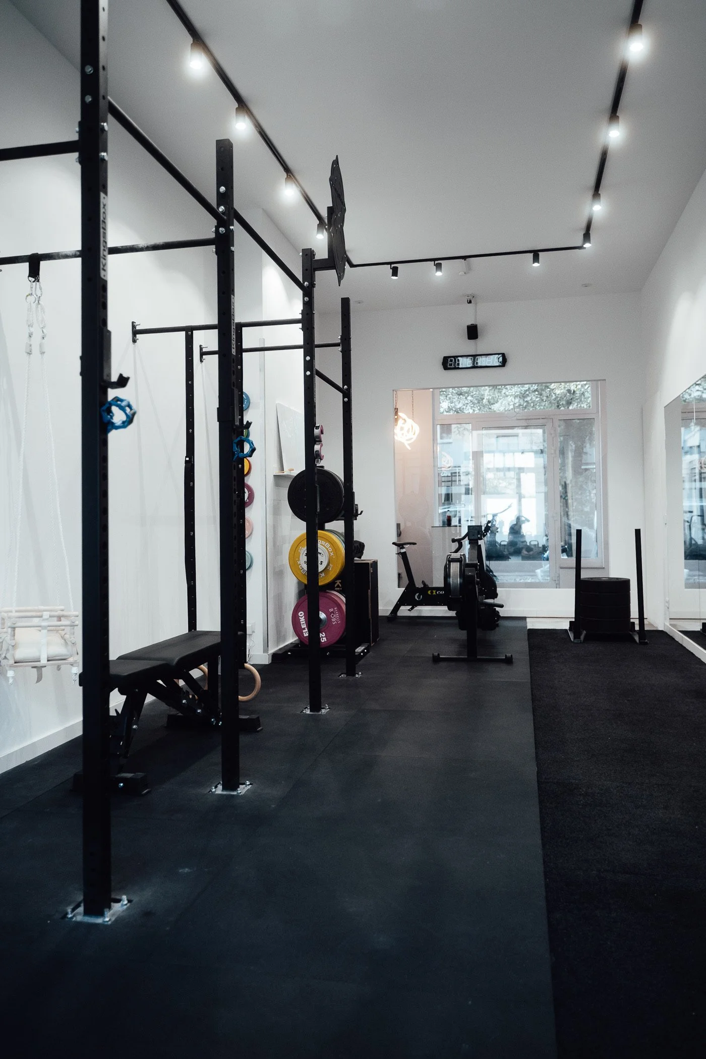 Leerer Fitnessraum mit Gymnastikgerät, Gewichten und Fahrrad, helle Wände, viele Fenster, dunkler Boden, Deckenbeleuchtung, Uhr und Thermometer an der Wand.