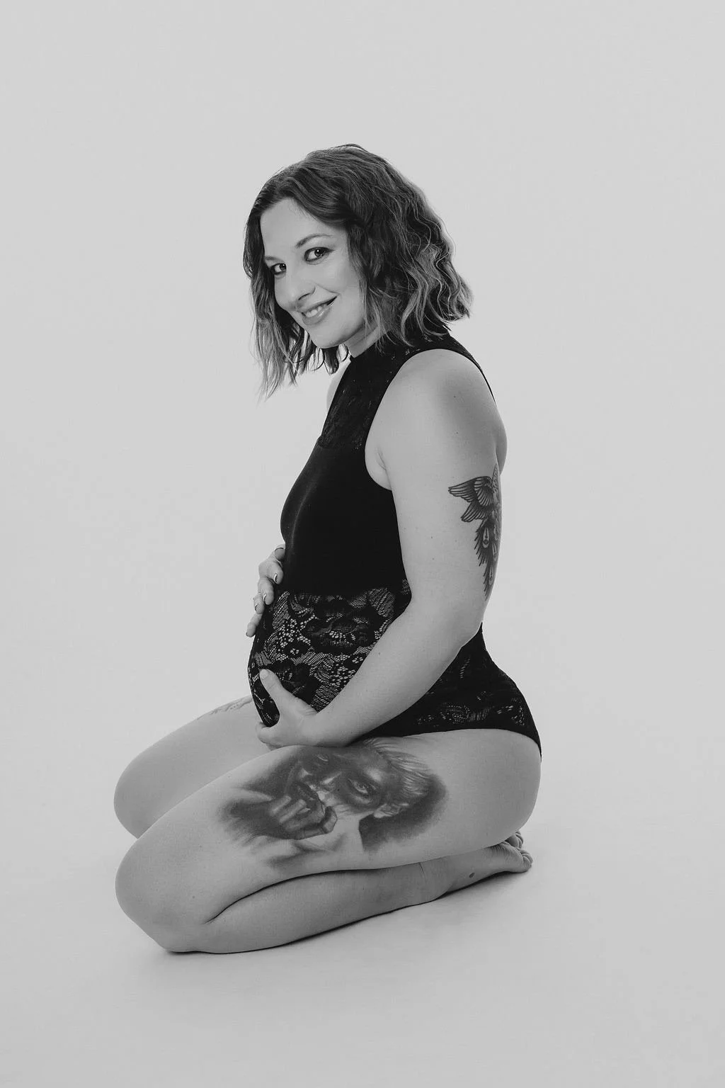 Schwangere Frau mit Tattoos, sitzend auf Knien, in schwarzer Lingerie, lächelnd, schwarze und weiße Fotografie.