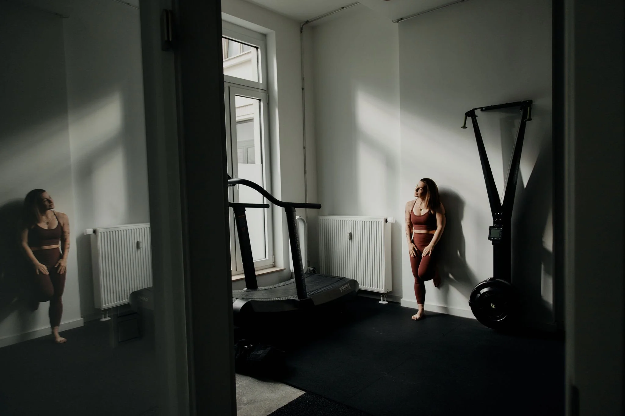 Frau in Mitarbeiterausfallsfitnessraum, die an der Wand lehnt, neben Laufband und Kraftgerät, mit Spiegel, weißen Wänden und Heizkörpern im Hintergrund.