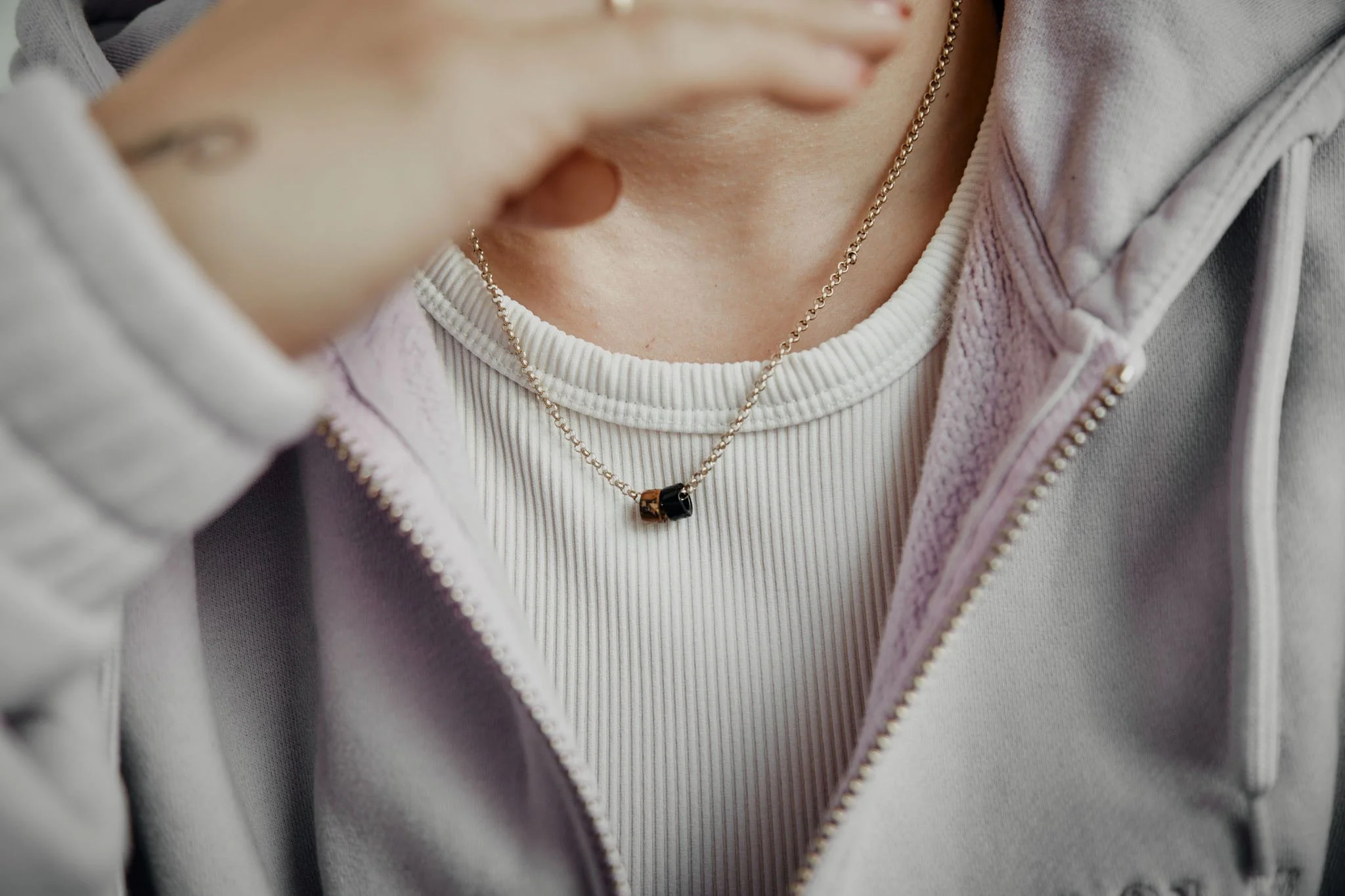 Nahaufnahme einer Frau, die eine goldene Halskette mit einem schwarzen Anhänger trägt, während sie eine graue Kapuzenjacke trägt.