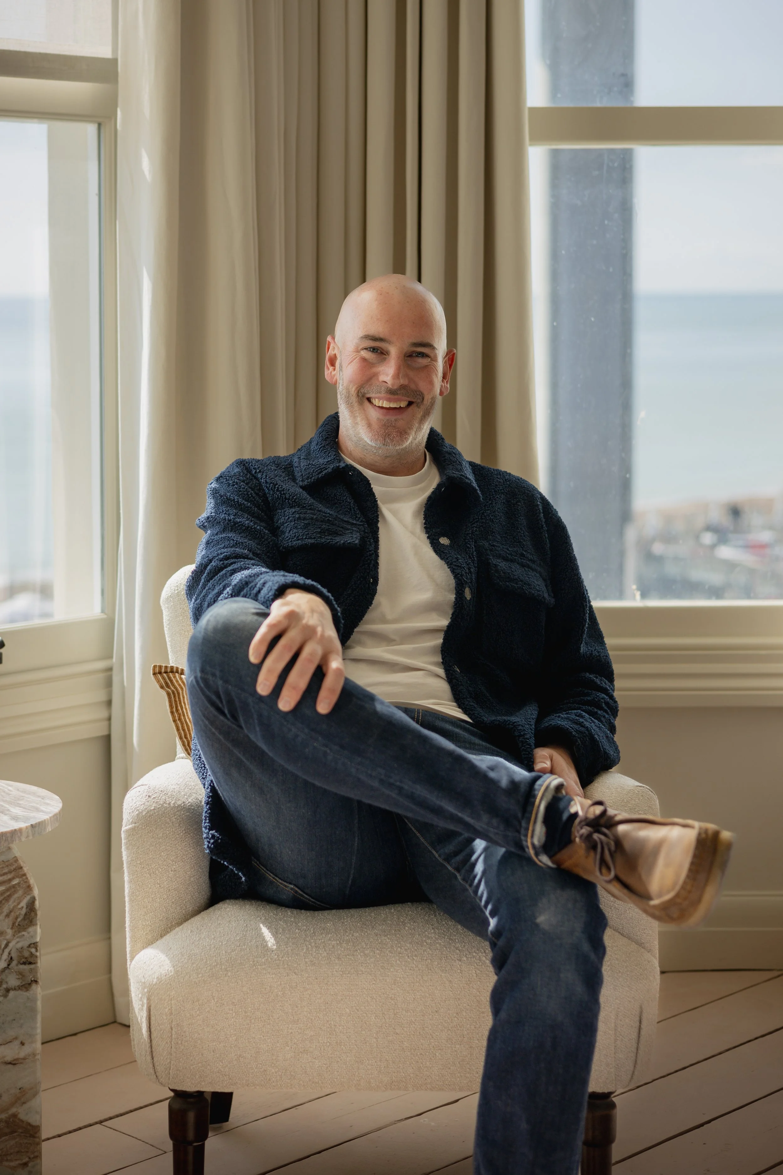 Smiling man sitting on a beige chair indoors near large windows, ocean view in background, dressed casually in a navy jacket, white t-shirt, jeans, and brown boots.