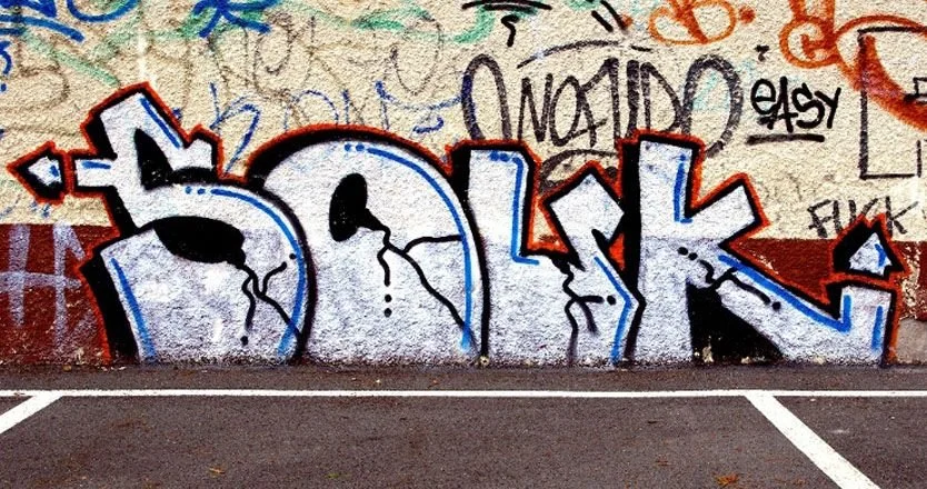 Graffiti coloré sur un mur, formant le mot "SOLK" avec des éclaboussures et des lignes noires et bleues, entouré d'autres inscriptions en arrière-plan. Graffiti a retirer