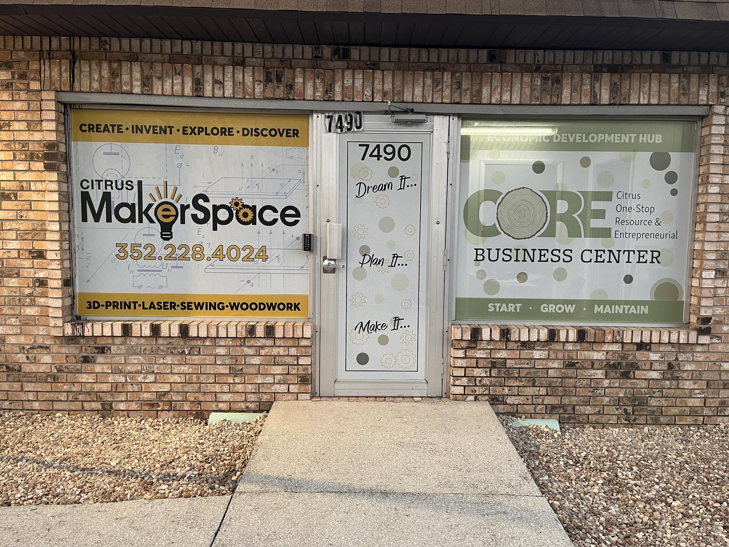 The front of a brick building with three large windows and a metal door in the center. The left window advertises Citrus MakerSpace with the words 'Create, Invent, Explore, Discover', a phone number, and services like 3D print, laser, sewing, and woo