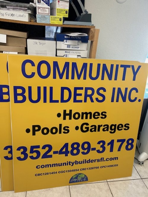 Sign for Community Builders Inc. advertising services for homes, pools, and garages, with a phone number and website, placed indoors near shelves with boxes and supplies.