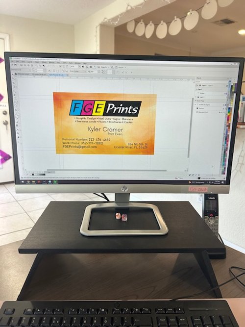 Computer monitor displaying a graphic design software with a branded card for FGE Prints showing contact information and logo, on a desk with a small pair of dice on a tray.