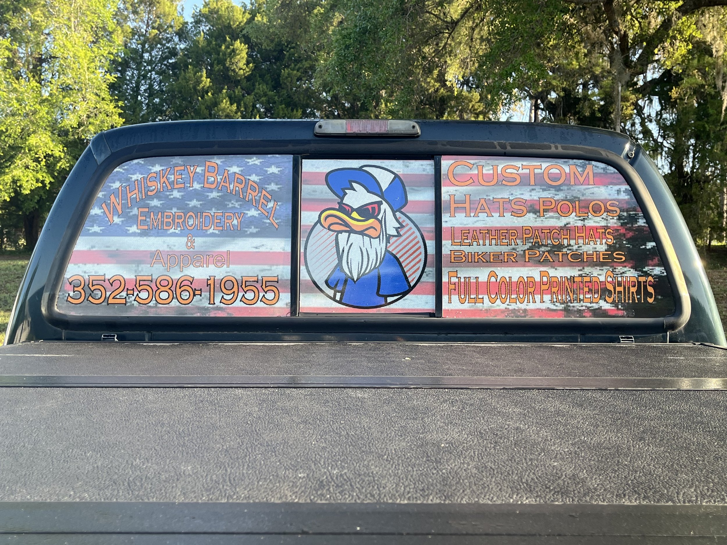 Decal advertising Whiskey Barrel Embroidery and Apparel on the back of a pickup truck with a cartoon eagle wearing a blue cap and jacket, against an American flag background.