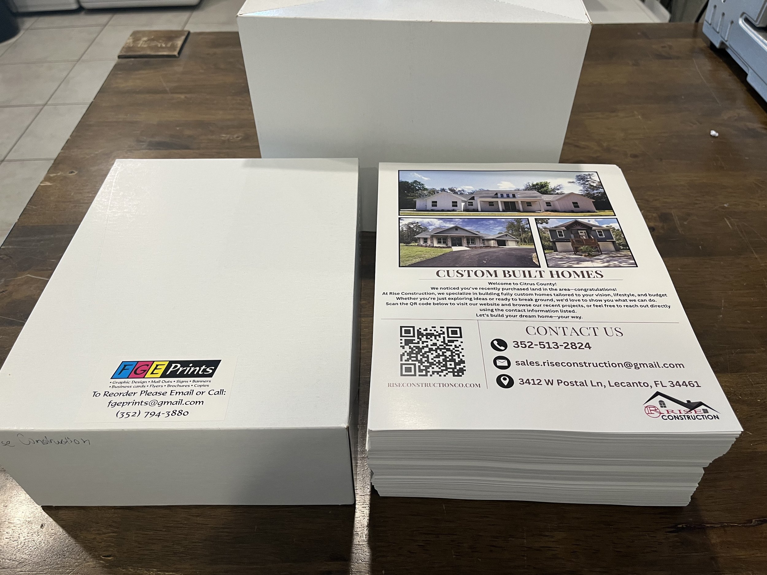 Stacks of printed flyers and boxes on a wooden table, advertising custom-built homes by Rise Construction with contact information, QR code, and images of different house designs.