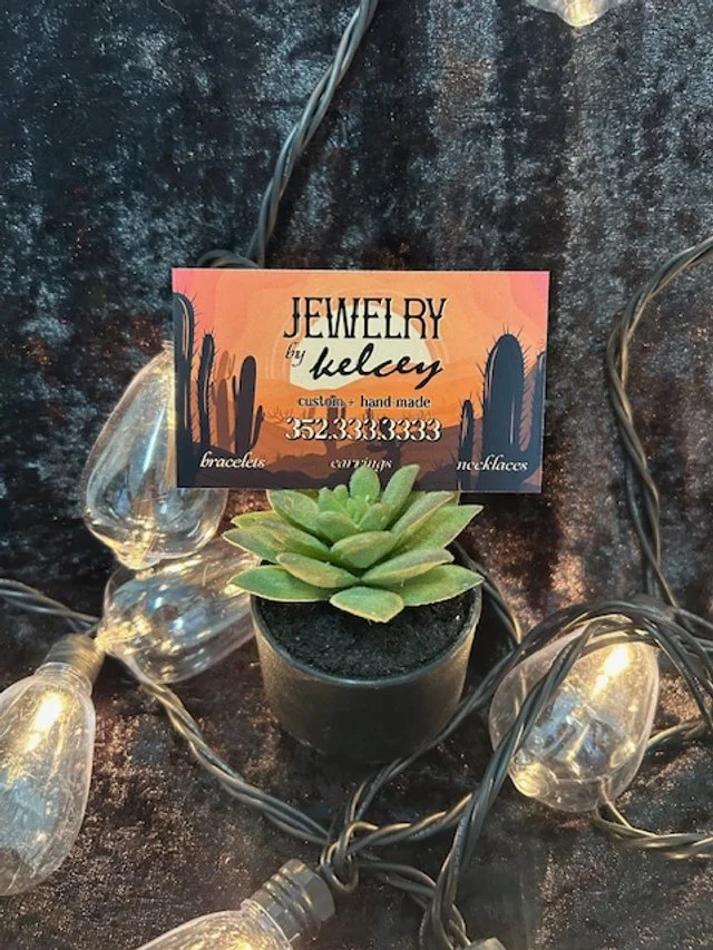 A small potted succulent plant surrounded by string lights on a dark surface, with a colorful business card for jewelry by Kelcy placed above the plant.