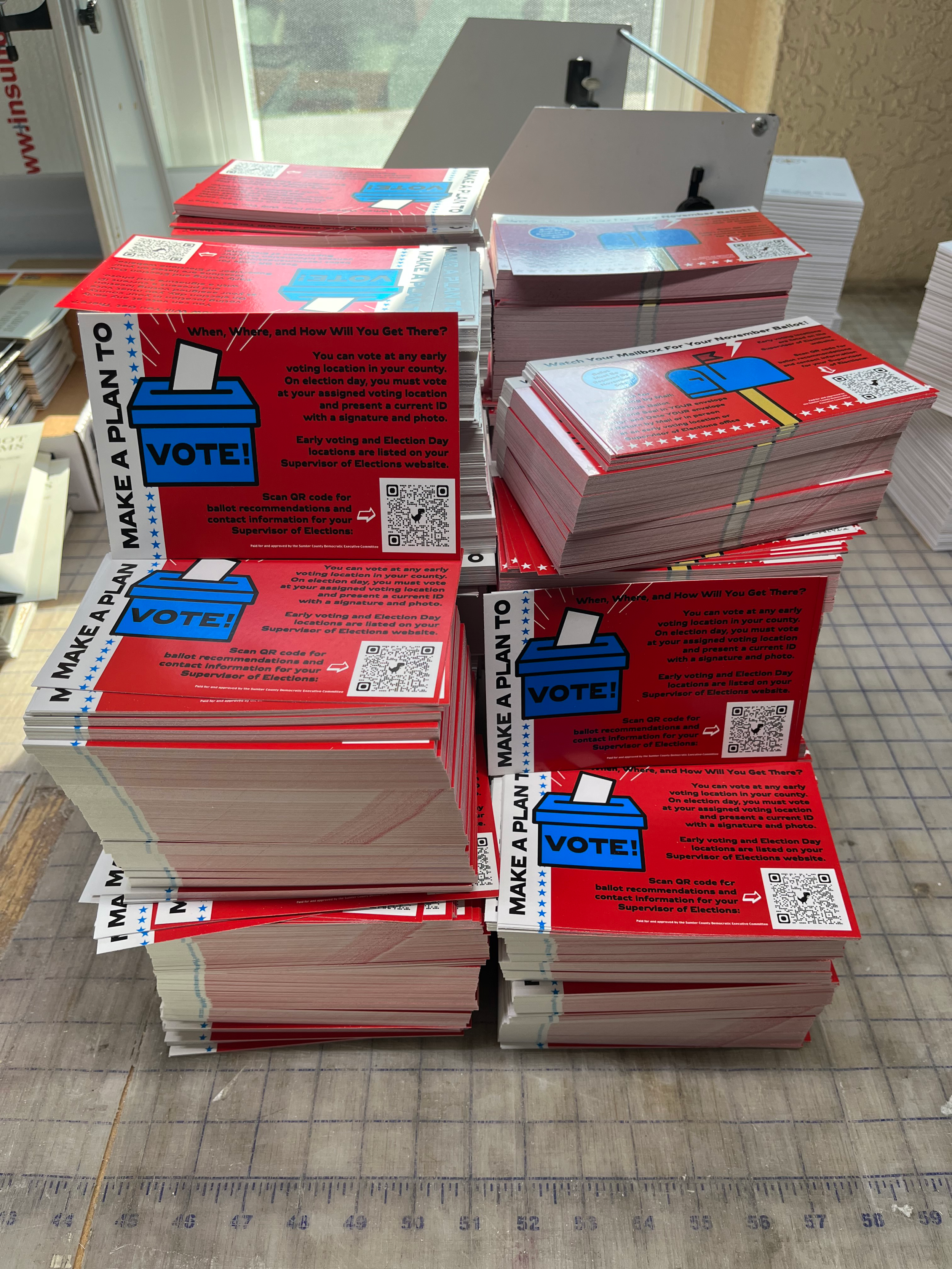 Stacks of red and white voting informational flyers with a blue ballot box icon and the word 'VOTE' on a table near a window.