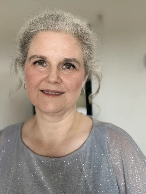 A close-up portrait of an older woman with gray hair, light skin, and a gentle smile, wearing a gray, shimmering top.