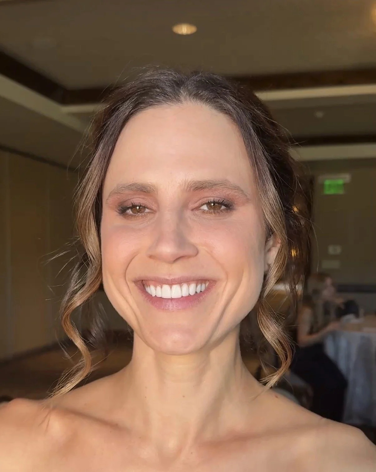 A smiling woman with wavy brown hair, wearing makeup, in a well-lit indoor setting with other people in the background.