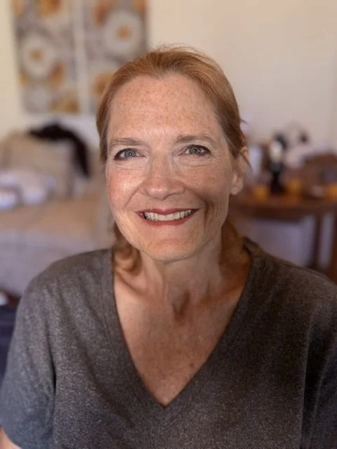 Smiling woman with red hair and freckles, wearing a dark gray shirt, in a cozy home setting.