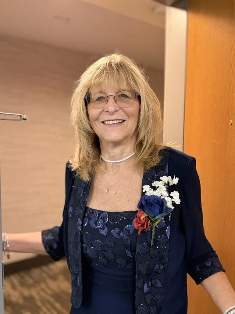 A smiling woman with glasses and blonde hair, wearing a dark blue dress and jacket, standing indoors with a floral corsage on her jacket.