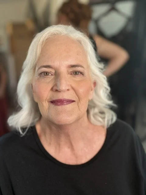 Close-up of an older woman with white hair smiling, wearing a black top, in an indoor setting.