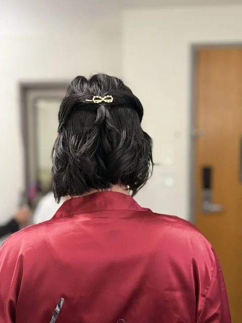 Back view of a woman with short, dark hair styled with a gold hair clip, wearing a red satin robe, in a room with a mirror and a door