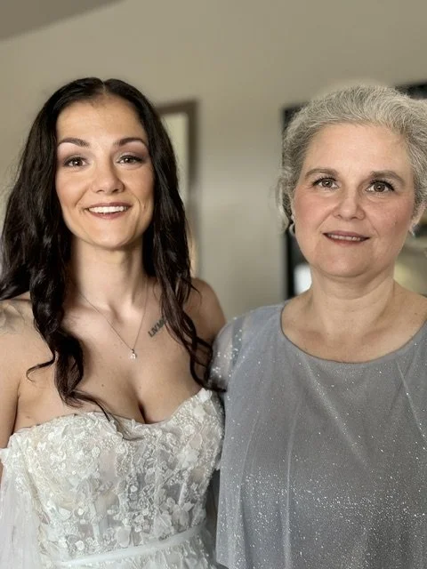 A young woman in a white dress smiling next to an older woman with gray hair in a gray dress, standing indoors in front of a beige wall.