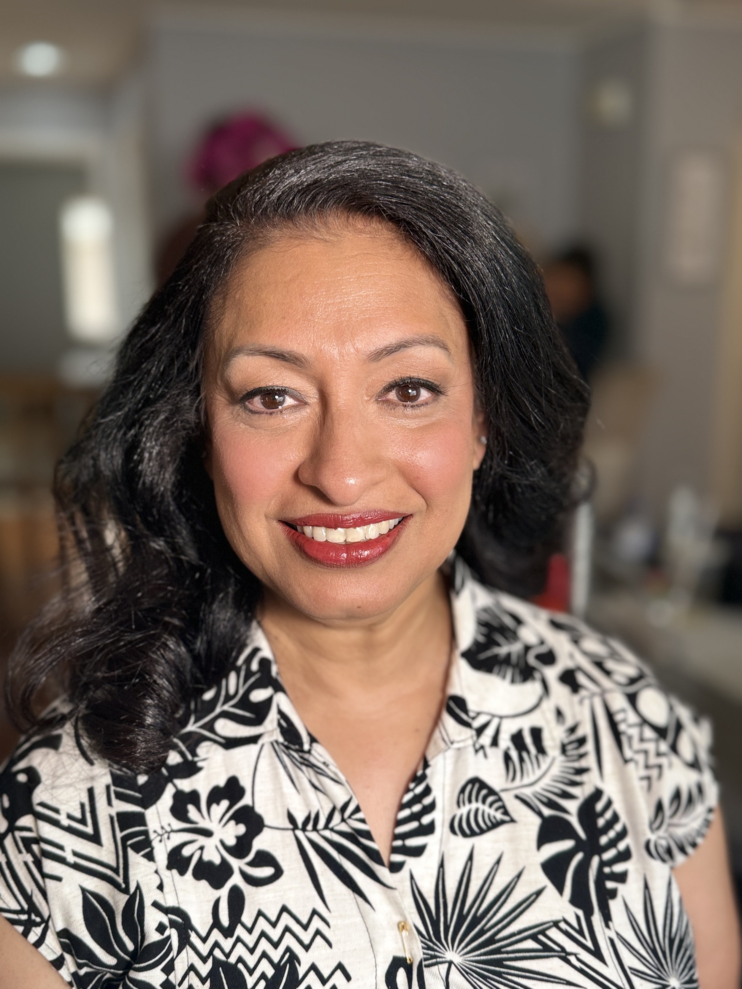Close-up of smiling woman with dark hair, wearing a black and white patterned shirt, in an indoor setting.