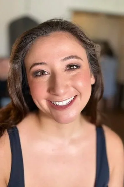 A young woman with brown hair and a big smile, wearing a dark blue sleeveless top, indoors with a blurred background.