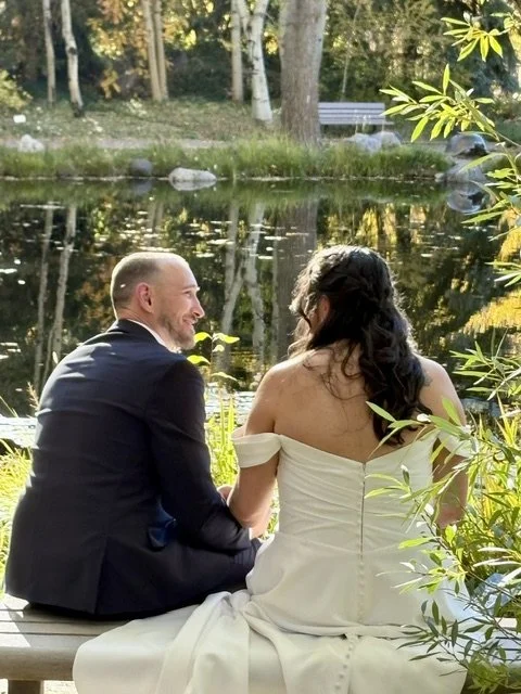 A newlywed couple sitting by a pond with trees and a park bench in the background, smiling at each other.
