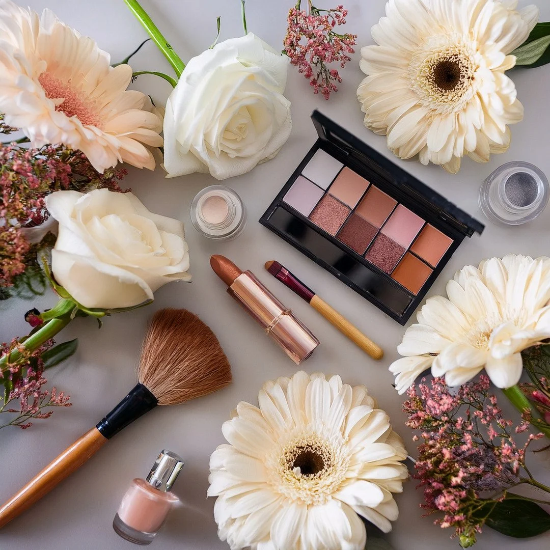 Makeup palette, brushes, lipstick, and nail polish surrounded by white and pink flowers on a white surface.