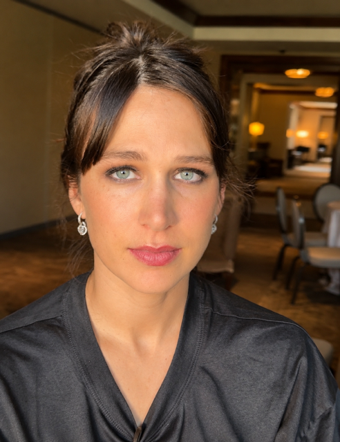 A close-up portrait of a woman with blue eyes, dark brown hair styled in an updo, wearing earrings and a black top, indoors with warm lighting and a blurred background of a dining area.