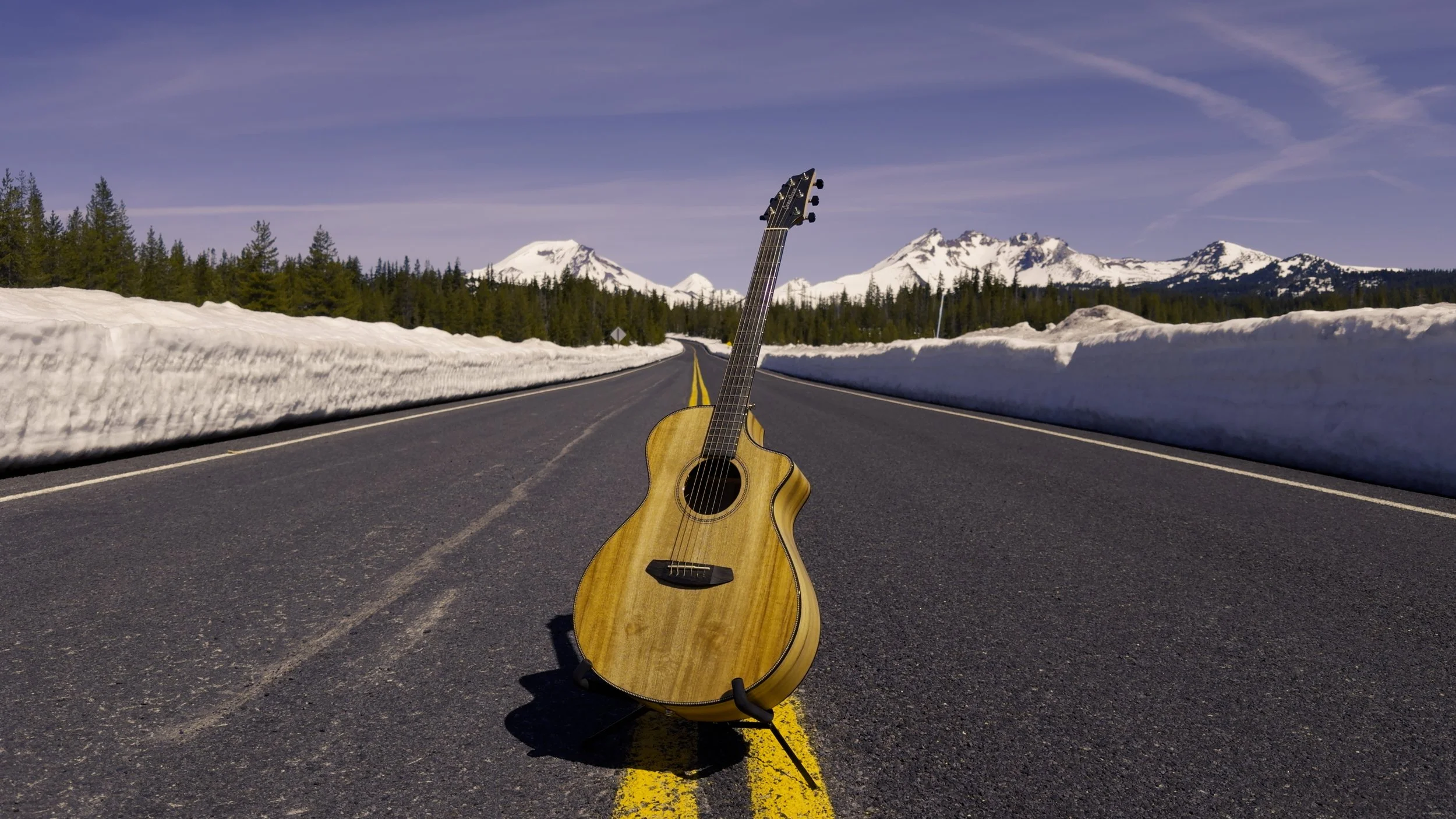 An acoustic guitar standing upright in the middle of a two-lane road with snow piled on either side, against a backdrop of mountains and a blue sky.